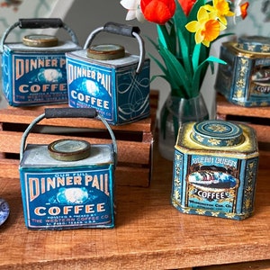 May include: A collection of vintage coffee tins in blue and gold with the words "Dinner pail coffee" and "Ricabi Queen Coffee". The tins are arranged on a wooden surface with a coffee grinder and a bouquet of yellow and red flowers.