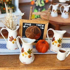 May include: Miniature ceramic pitchers with autumn leaf designs, a small pumpkin, and a tiny cup of tea are arranged on a table. A sign reads "ONE INCH" next to a penny. The scene evokes a cozy fall theme.