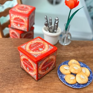 May include: Miniature red Amaretti biscuits tins with gold lettering and a plate of almond cookies. A small vase with a single orange flower and a small white pot with silver utensils are also visible.