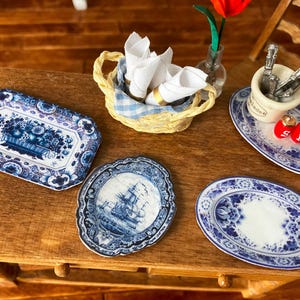 May include: Miniature blue and white porcelain plates and serving dishes. The set includes a rectangular tray with a floral design, a round plate with a ship, and oval plates. A basket of napkins, silverware, and salt and pepper shakers complete the dining set.