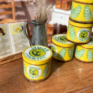 May include: A miniature kitchen scene with a cookbook, a metal pitcher with dried flowers, and several yellow tins of "Ginger Cookies." The tins have a green and white design. The scene is set on a wooden surface.