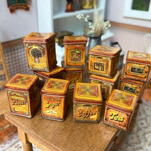 May include: A collection of miniature vintage-style tin boxes, labelled for flour, sugar, coffee, and tea. The boxes are primarily yellow and brown with decorative floral and wheat designs. They are arranged on a small wooden table.