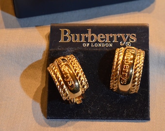 May include: A pair of gold-tone hoop earrings with the Burberrys of London logo engraved on the front. The earrings are displayed on a black velvet background.