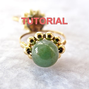 May include: A gold ring with a green jade stone surrounded by gold beads. The ring is on a white fuzzy surface.