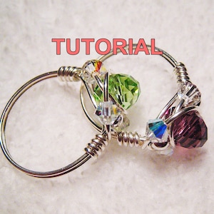 May include: A silver wire ring with a green, purple, and clear crystal design. The ring is wrapped with silver wire and has a beaded design. The ring is a handmade piece of jewelry.