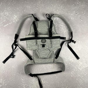 May include: A gray baby carrier with black straps and buckles. The carrier has a patterned design and a circular logo on the front. The shoulder straps are padded for comfort.