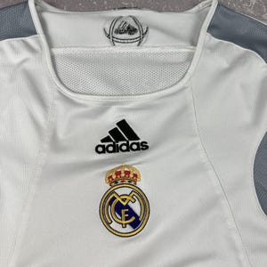 Puede incluir: Una camiseta deportiva blanca con paneles de malla gris en los hombros. El pecho muestra un logotipo negro de Adidas sobre el escudo del Real Madrid, que incluye una corona dorada, detalles en azul y oro, y las letras MCF en el centro.