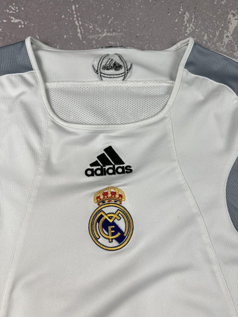 Puede incluir: Una camiseta deportiva blanca con paneles de malla gris en los hombros. El pecho muestra un logotipo negro de Adidas sobre el escudo del Real Madrid, que incluye una corona dorada, detalles en azul y oro, y las letras MCF en el centro.