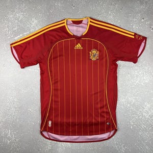May include: A red short-sleeve soccer jersey featuring thin vertical yellow stripes, a yellow Adidas logo on the chest, and the Spanish national team crest. The shirt includes yellow shoulder stripes and a round neckline with a light pink interior.