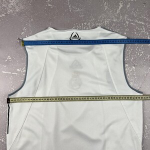 Puede incluir: Una camiseta deportiva sin mangas de color blanco con ribetes grises y el logotipo de Adidas impreso en el cuello. Una cinta m&eacute;trica azul sobre los hombros muestra 48 cm, y una cinta m&eacute;trica amarilla sobre el pecho muestra 53 cm.