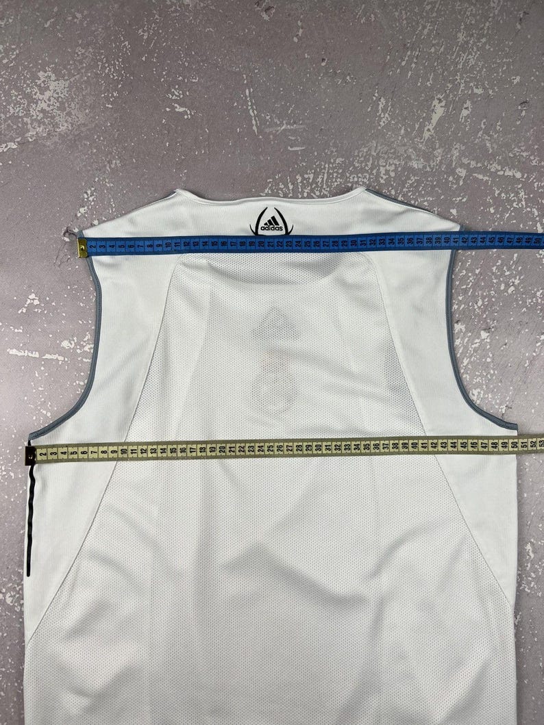 Puede incluir: Una camiseta deportiva sin mangas de color blanco con ribetes grises y el logotipo de Adidas impreso en el cuello. Una cinta m&eacute;trica azul sobre los hombros muestra 48 cm, y una cinta m&eacute;trica amarilla sobre el pecho muestra 53 cm.