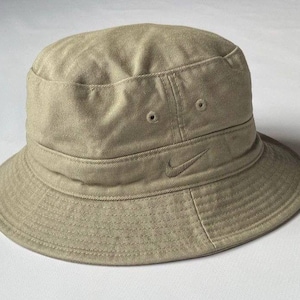 Op de afbeelding: Een beige bucket hat met een platte bovenkant en een naar beneden lopende rand. De hoed is gemaakt van geweven stof en is voorzien van een klein, ton-sur-ton geborduurd Nike swoosh-logo op het voorpaneel, samen met twee gestikte ventilatiegaatjes.