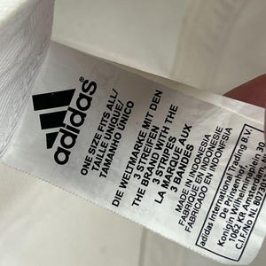 May include: A white fabric care label featuring the black Adidas logo and text in multiple languages including One Size Fits All, The Brand With The 3 Stripes, Made In Indonesia, and the address for Adidas International Trading B.V. in Amsterdam.