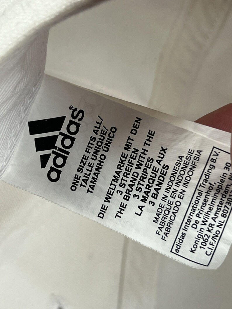 May include: A white fabric care label featuring the black Adidas logo and text in multiple languages including One Size Fits All, The Brand With The 3 Stripes, Made In Indonesia, and the address for Adidas International Trading B.V. in Amsterdam.