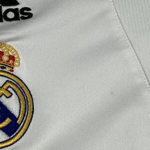 Puede incluir: Una camiseta deportiva blanca con el logotipo de Adidas en negro en la parte superior y el escudo del Real Madrid bordado debajo. El escudo muestra una corona dorada, un borde circular azul y dorado, y una franja diagonal azul sobre fondo blanco.