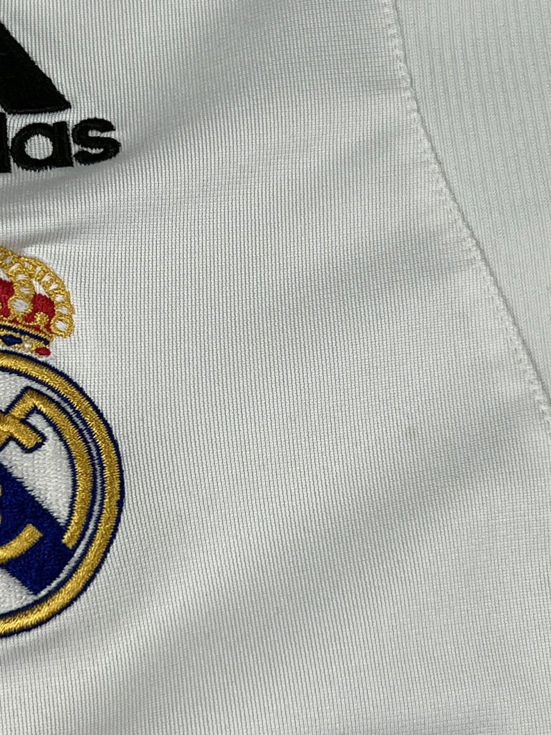 Puede incluir: Una camiseta deportiva blanca con el logotipo de Adidas en negro en la parte superior y el escudo del Real Madrid bordado debajo. El escudo muestra una corona dorada, un borde circular azul y dorado, y una franja diagonal azul sobre fondo blanco.