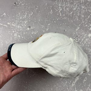 May include: A white baseball cap featuring a dark blue trim on the edge of the brim with the text Real Madrid C.F. printed in white. The cap includes a circular embroidered emblem on the front panel and ventilation eyelets on the crown.