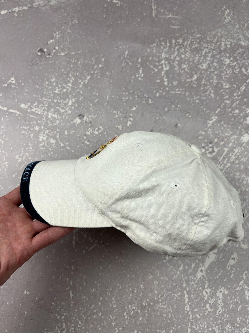 May include: A white baseball cap featuring a dark blue trim on the edge of the brim with the text Real Madrid C.F. printed in white. The cap includes a circular embroidered emblem on the front panel and ventilation eyelets on the crown.