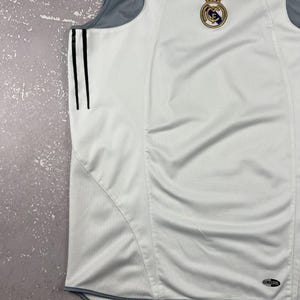 Puede incluir: Una camiseta deportiva sin mangas de color blanco con paneles laterales de malla gris y tres rayas verticales negras en el lateral. La prenda muestra el logotipo de Adidas y el escudo del Real Madrid en el pecho, con una etiqueta Climacool abajo.