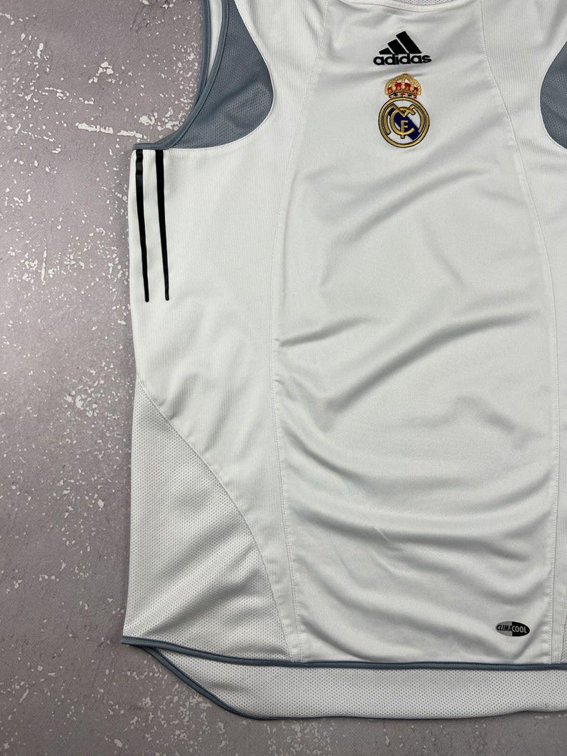 Puede incluir: Una camiseta deportiva sin mangas de color blanco con paneles laterales de malla gris y tres rayas verticales negras en el lateral. La prenda muestra el logotipo de Adidas y el escudo del Real Madrid en el pecho, con una etiqueta Climacool abajo.