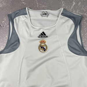 Puede incluir: Una camiseta deportiva sin mangas de color blanco con paneles de malla gris en los hombros. El pecho muestra un logotipo negro de Adidas sobre el escudo del club Real Madrid. El interior del cuello incluye un peque&ntilde;o gr&aacute;fico circular.