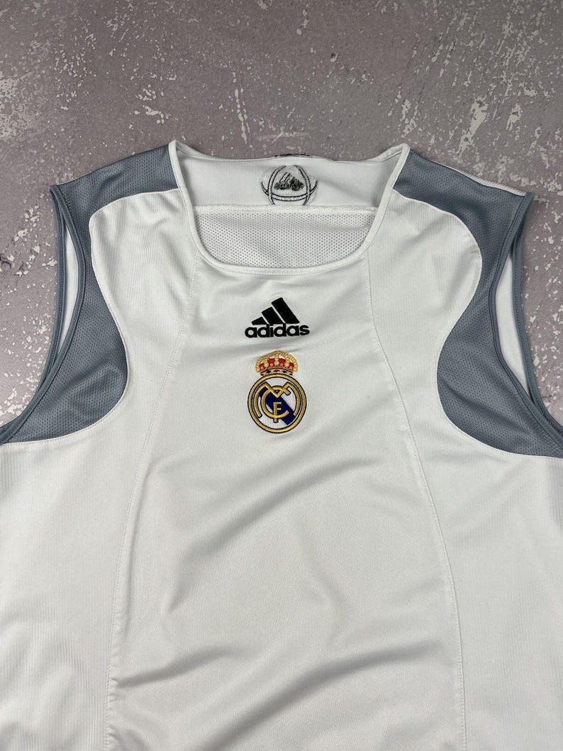 Puede incluir: Una camiseta deportiva sin mangas de color blanco con paneles de malla gris en los hombros. El pecho muestra un logotipo negro de Adidas sobre el escudo del club Real Madrid. El interior del cuello incluye un peque&ntilde;o gr&aacute;fico circular.