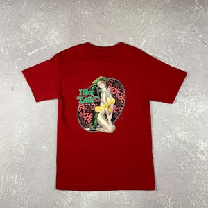 May include: Red t-shirt with a graphic on the back. The graphic shows a woman with flowers, surrounded by a floral design. The text "I Got Lei'd" is printed above the image in green and yellow.