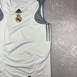Puede incluir: Una camiseta deportiva sin mangas de color blanco con paneles de malla gris en los hombros y los costados. La prenda muestra un logotipo de Adidas negro, el escudo del Real Madrid y dos rayas verticales negras en el lateral. Etiqueta Climacool en el dobladillo.