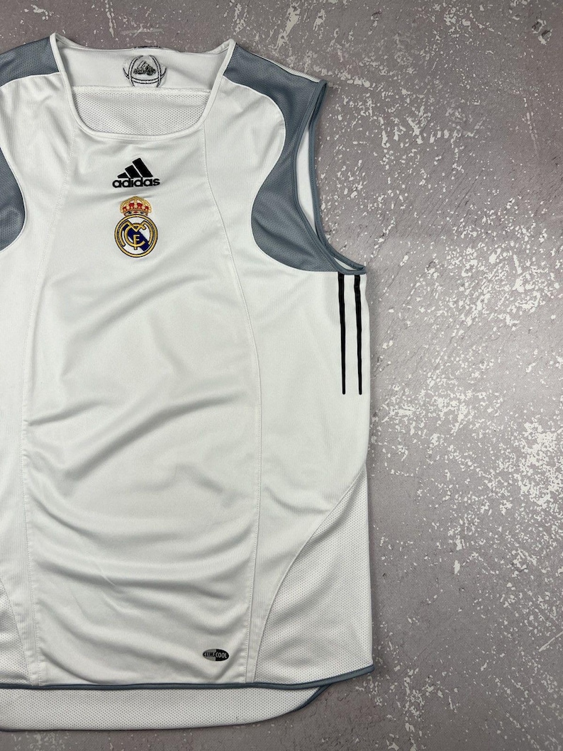 Puede incluir: Una camiseta deportiva sin mangas de color blanco con paneles de malla gris en los hombros y los costados. La prenda muestra un logotipo de Adidas negro, el escudo del Real Madrid y dos rayas verticales negras en el lateral. Etiqueta Climacool en el dobladillo.