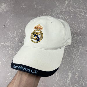 May include: An off-white baseball cap featuring the Real Madrid C.F. logo embroidered on the front crown in blue, gold, and white. The brim has a dark navy blue edge with the text Real Madrid C.F. embroidered in light blue lettering.