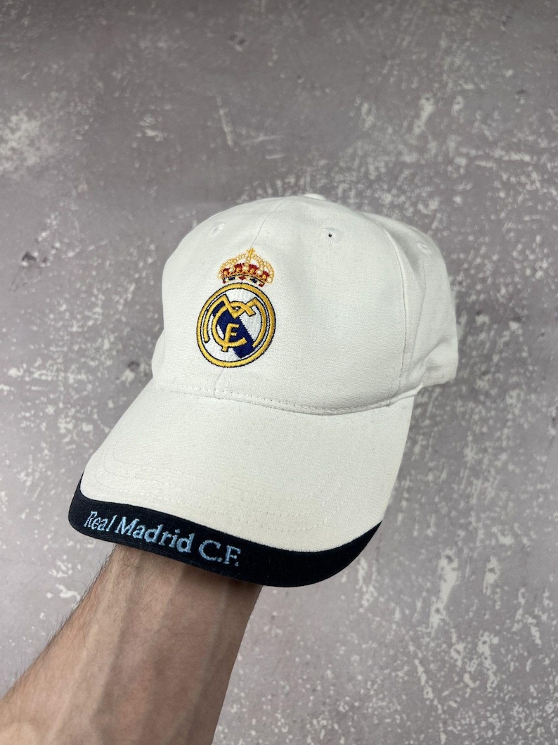 May include: An off-white baseball cap featuring the Real Madrid C.F. logo embroidered on the front crown in blue, gold, and white. The brim has a dark navy blue edge with the text Real Madrid C.F. embroidered in light blue lettering.