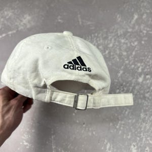 May include: An off-white baseball cap featuring a black embroidered Adidas logo above the back opening. The cap includes a fabric strap with a silver-toned metal buckle for adjustment and stitched eyelets on the crown panels.