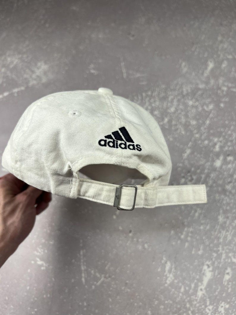May include: An off-white baseball cap featuring a black embroidered Adidas logo above the back opening. The cap includes a fabric strap with a silver-toned metal buckle for adjustment and stitched eyelets on the crown panels.