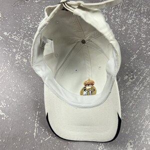 May include: A white baseball cap viewed from the inside, featuring a metal adjustable strap at the back and a Real Madrid club crest embroidered in gold, blue, and red on the interior front panel. The brim has a black trim along the edge.