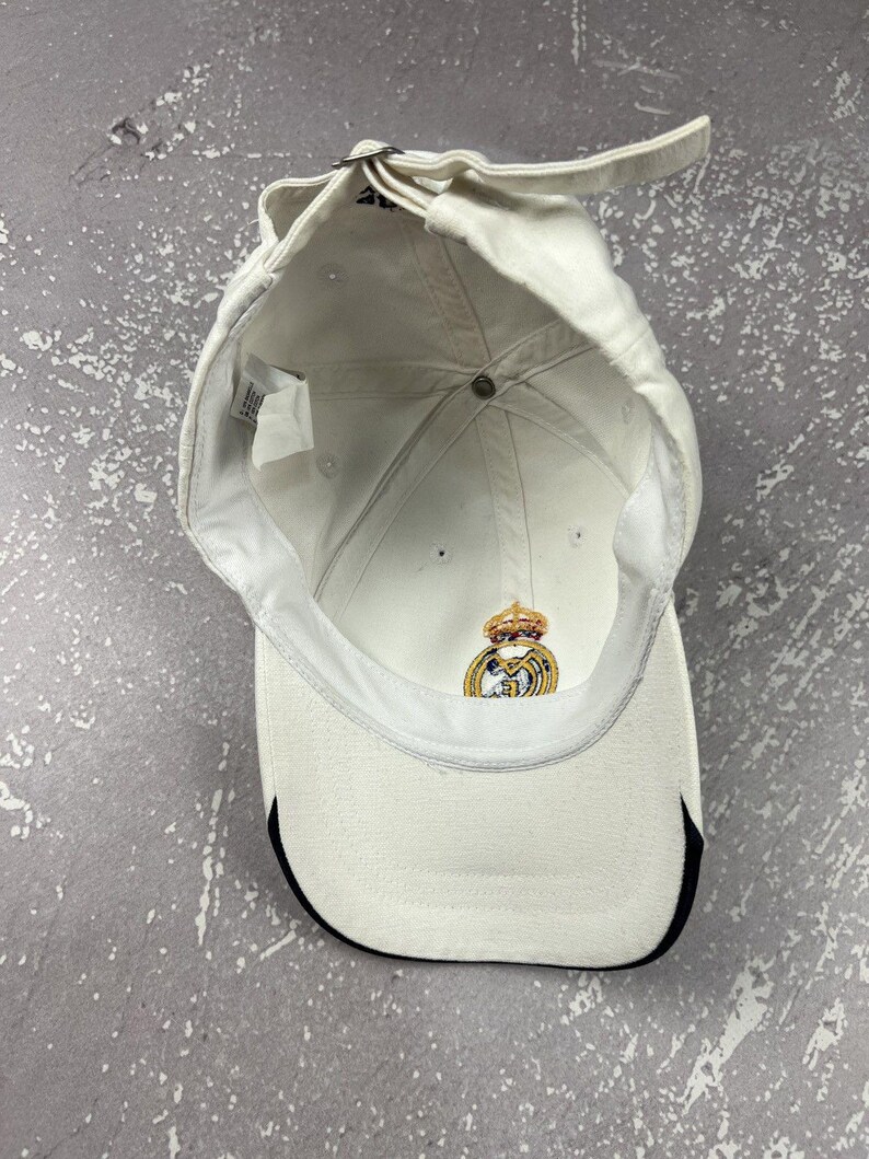 May include: A white baseball cap viewed from the inside, featuring a metal adjustable strap at the back and a Real Madrid club crest embroidered in gold, blue, and red on the interior front panel. The brim has a black trim along the edge.
