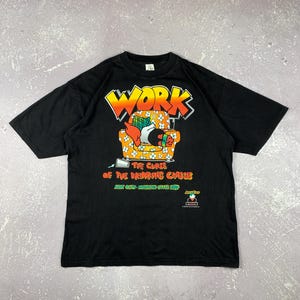 May include: Black t-shirt with a graphic design featuring the word "WORK" in orange and yellow, above a cartoon image of a person on a floral couch. The text "THE CURSE OF THE DRINKING CLASS" is below the couch.