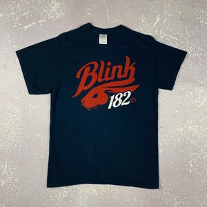 May include: Navy blue t-shirt featuring the band name "Blink 182" in red script with a red rabbit graphic and the number 182 in white. The shirt is a classic crew neck style.