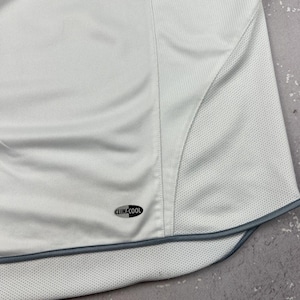Puede incluir: Una camiseta deportiva blanca con un panel frontal liso y paneles laterales de tejido de malla transpirable. La prenda incluye un borde gris oscuro a lo largo del dobladillo curvo y un peque&ntilde;o logotipo ovalado negro con el texto CLIMACOOL.
