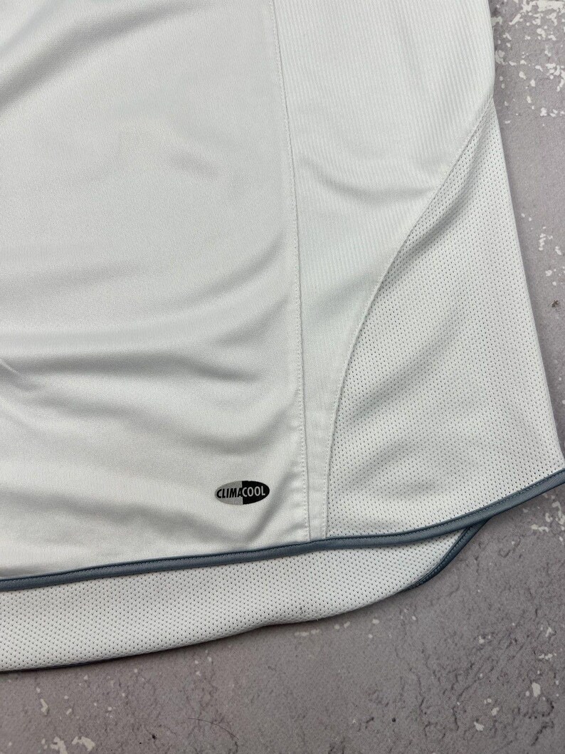 Puede incluir: Una camiseta deportiva blanca con un panel frontal liso y paneles laterales de tejido de malla transpirable. La prenda incluye un borde gris oscuro a lo largo del dobladillo curvo y un peque&ntilde;o logotipo ovalado negro con el texto CLIMACOOL.