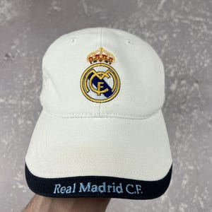 May include: A white baseball cap featuring the Real Madrid C.F. crest embroidered in blue, gold, and red on the front panel. The brim has a dark navy blue edge with the text Real Madrid C.F. embroidered in light blue lettering across the front.