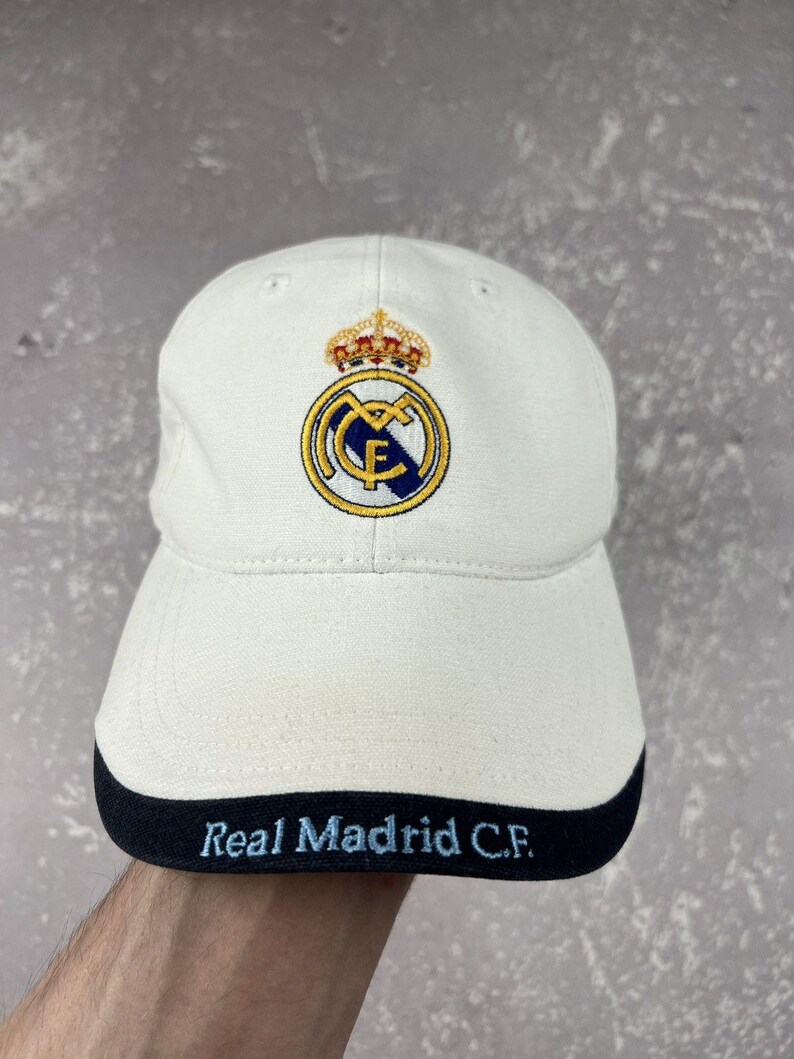 May include: A white baseball cap featuring the Real Madrid C.F. crest embroidered in blue, gold, and red on the front panel. The brim has a dark navy blue edge with the text Real Madrid C.F. embroidered in light blue lettering across the front.