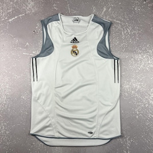 Puede incluir: Una camiseta deportiva sin mangas de color blanco con paneles grises en los hombros y dos finas rayas verticales negras a cada lado. La prenda muestra el logo de Adidas en negro y el escudo del Real Madrid en el pecho, con un emblema Climacool.