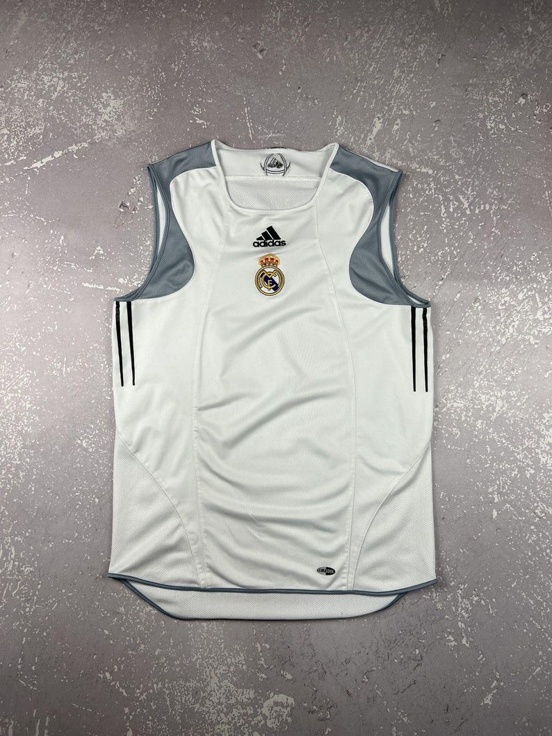 Puede incluir: Una camiseta deportiva sin mangas de color blanco con paneles grises en los hombros y dos finas rayas verticales negras a cada lado. La prenda muestra el logo de Adidas en negro y el escudo del Real Madrid en el pecho, con un emblema Climacool.