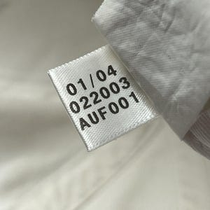 May include: A white fabric garment tag with black printed text. The tag displays the numbers 01/04 and 022003, followed by the alphanumeric code AUF001. The tag is attached to a white textile surface with a visible woven texture and stitching.