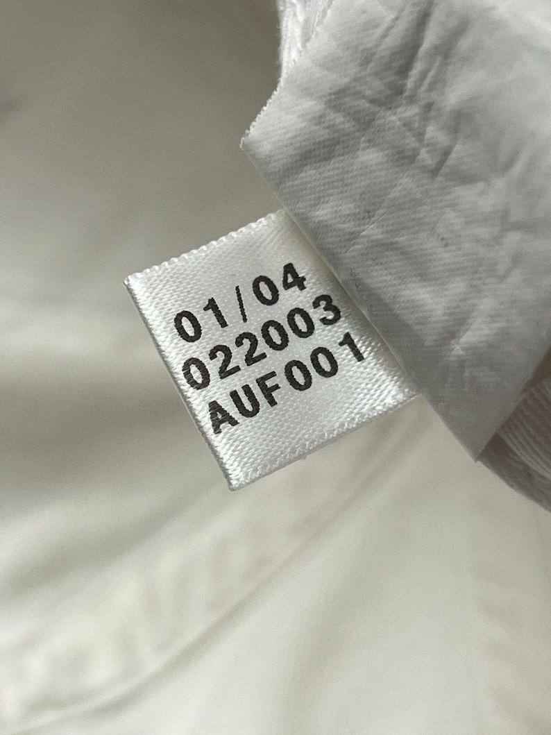 May include: A white fabric garment tag with black printed text. The tag displays the numbers 01/04 and 022003, followed by the alphanumeric code AUF001. The tag is attached to a white textile surface with a visible woven texture and stitching.