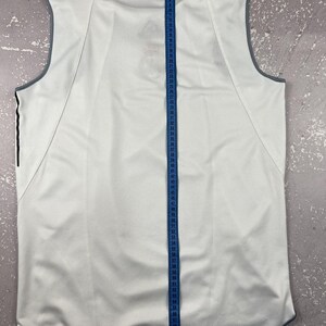 Puede incluir: Una camiseta deportiva sin mangas de color blanco con ribetes grises y el logotipo de Adidas en el cuello. Una cinta m&eacute;trica azul est&aacute; colocada verticalmente en el centro, mostrando una medida de longitud superior a 165 cm.