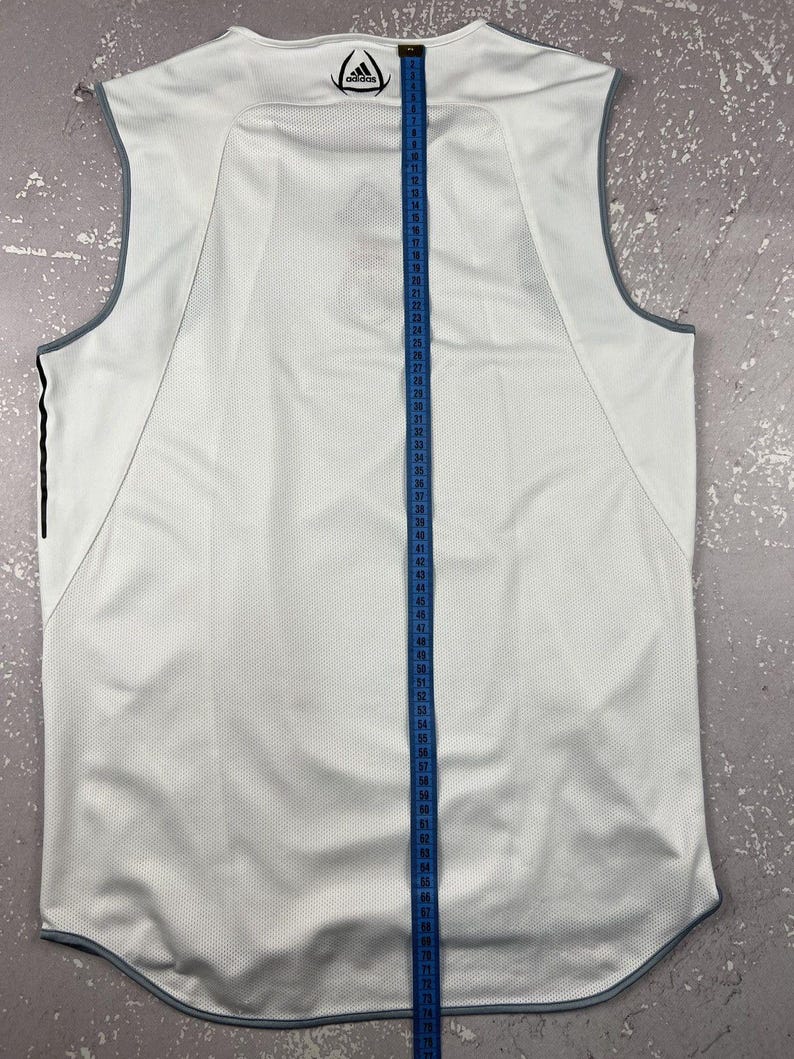 Puede incluir: Una camiseta deportiva sin mangas de color blanco con ribetes grises y el logotipo de Adidas en el cuello. Una cinta m&eacute;trica azul est&aacute; colocada verticalmente en el centro, mostrando una medida de longitud superior a 165 cm.