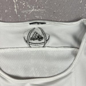 Puede incluir: Una camiseta deportiva blanca con cuello redondo y un panel de malla. Un parche con un logotipo circular bordado en blanco y negro se sit&uacute;a en el centro del cuello, y un logotipo de tres rayas negras es visible en el borde inferior.