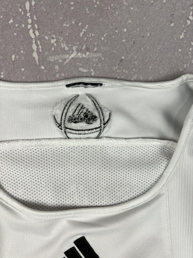 Puede incluir: Una camiseta deportiva blanca con cuello redondo y un panel de malla. Un parche con un logotipo circular bordado en blanco y negro se sit&uacute;a en el centro del cuello, y un logotipo de tres rayas negras es visible en el borde inferior.