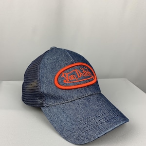 May include: A denim trucker hat featuring a dark blue mesh back and a solid denim front panel. An oval orange patch is embroidered on the front with the text Von Dutch in orange lettering. The hat has a curved brim and a structured crown design.